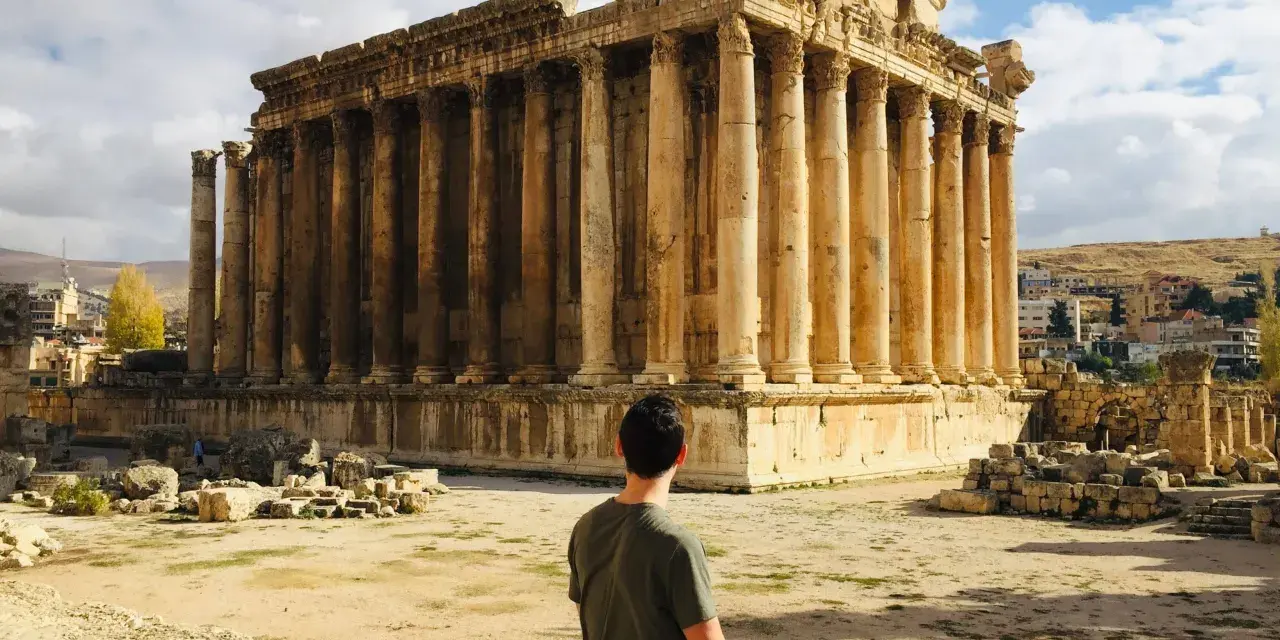 A man sits contemplating the grandeur of Roman ruins in Lebanon, bathed in sunlight under a cloudy sky.