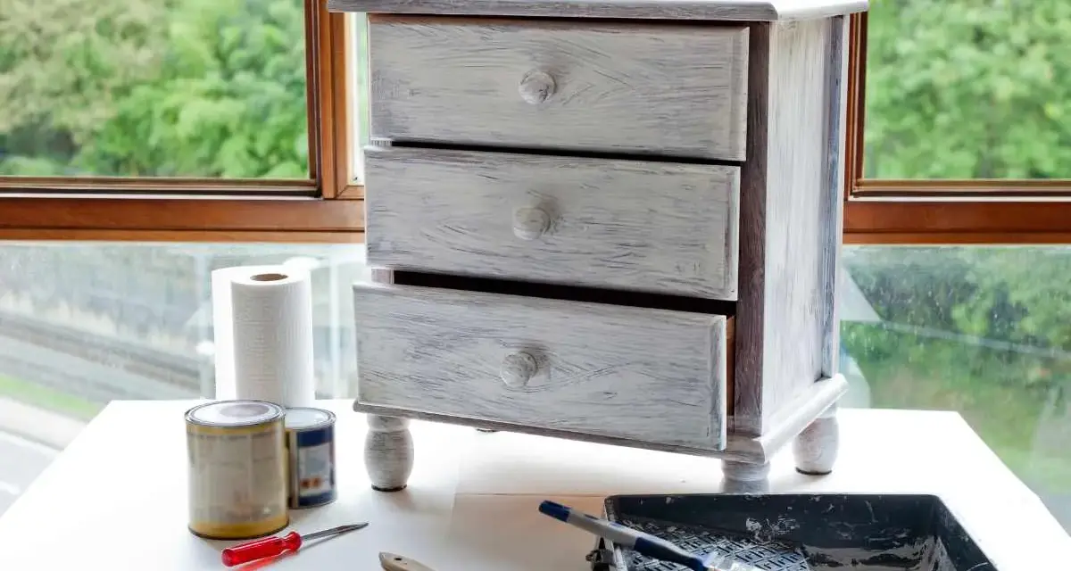 Mueble de tres cajones con acabado desgastado, listo para pintar muebles oscuros. Pinceles, rodillo y latas de pintura a su lado.