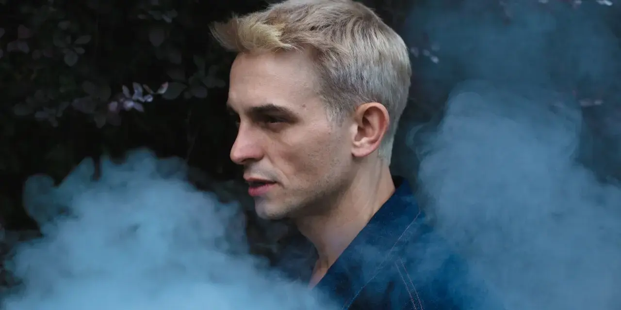 Vito Bambino w dymie, zapowiada nową płytę. Artysta w denimowej kurtce, z blond włosami, na tle ciemnej roślinności.