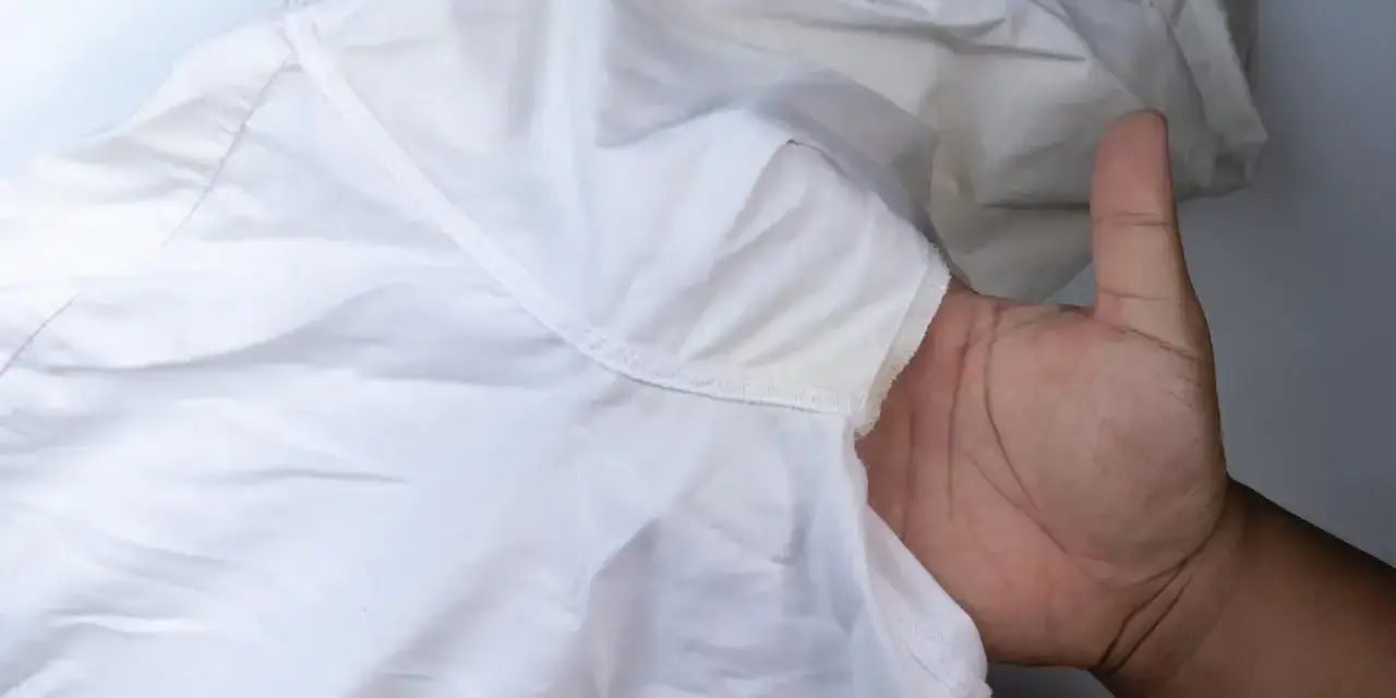 Biała koszula z widocznymi żółte plamy pod pachami. Dłoń trzyma materiał, ukazując problem.
