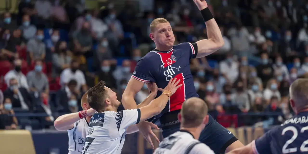 Dainis Kristopans w skoku rzuca piłką do bramki, otoczony przez obrońców.