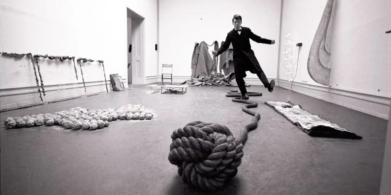 Un homme en costume, peut-être Lawrence Weiner, danse au milieu d'une installation artistique avec des cordes et des objets.