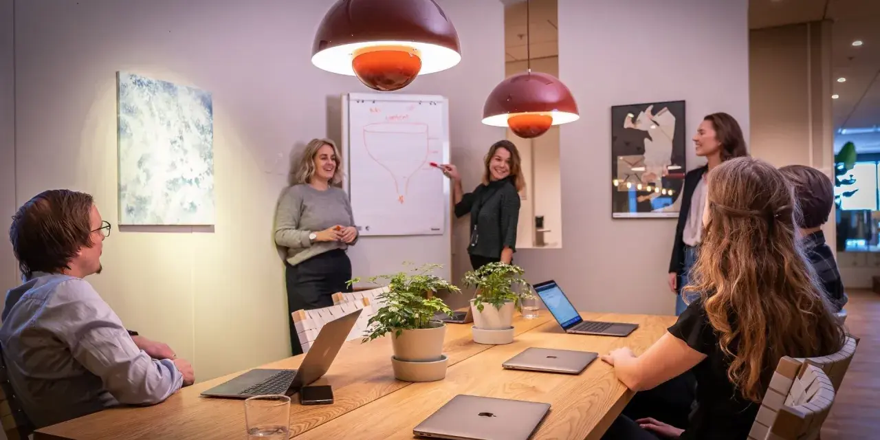 Ett team diskuterar strategier vid ett konferensbord med flera laptops. En kvinna pekar på en trattmodell på en whiteboard. Ups datorn är en del av deras effektiva arbetsflöde.