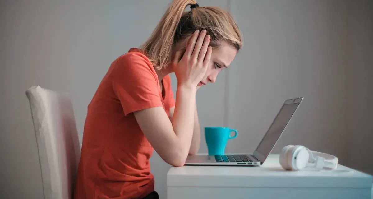 Zmartwiona kobieta przy laptopie, zastanawia się, czy porzucenie pracy wlicza się do emerytury. Na biurku kubek i słuchawki.
