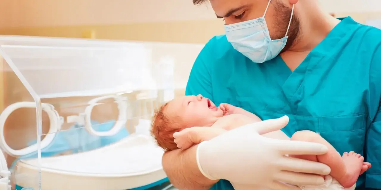 Lekarz w maseczce i rękawiczkach trzyma noworodka. To przykład troski w neonatologii, która zajmuje się opieką nad noworodkami.