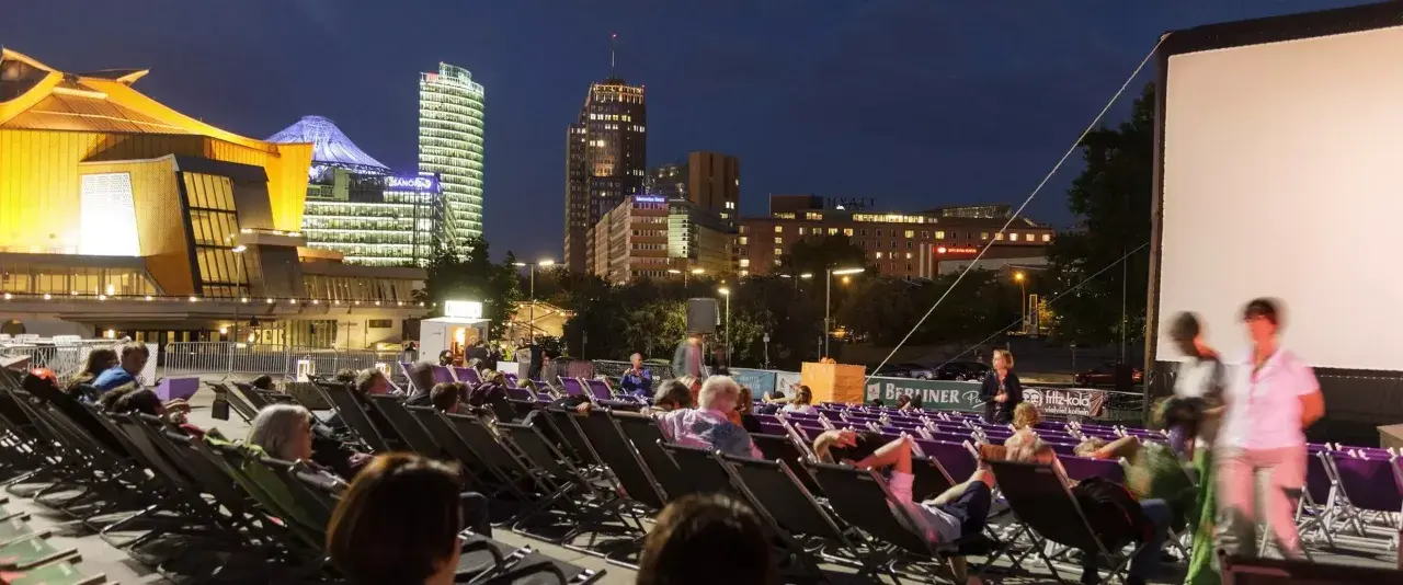 Leute entspannen sich in Liegestühlen beim **Sommerkino am Potsdamer Platz** vor einer leuchtenden Skyline.