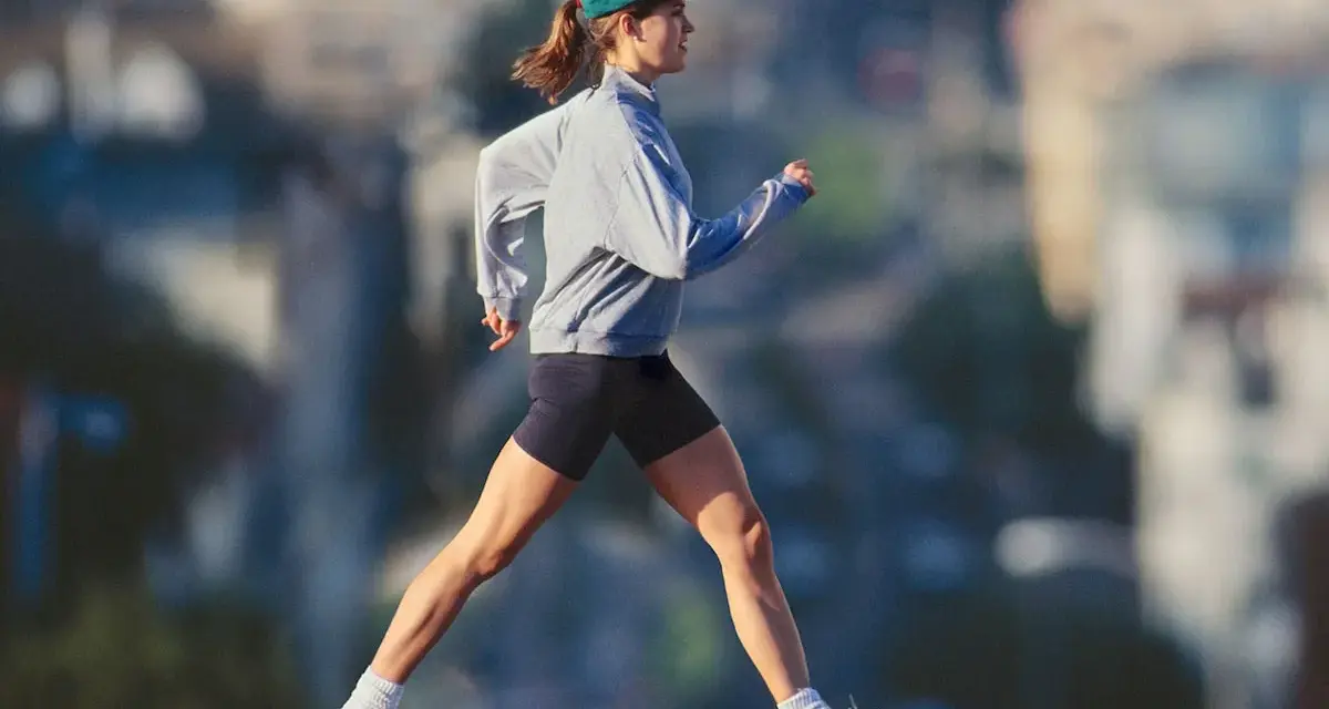 Mujer con gorra y sudadera gris corriendo al aire libre, disfrutando de un paseo después de comer para mantenerse activa.