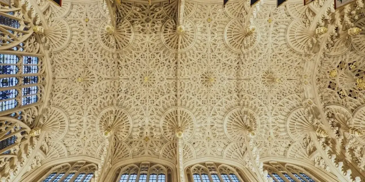 Intricate fan vaulting of a grand hall, reminiscent of the detailed ceilings found in abbey graves, adorned with flags.