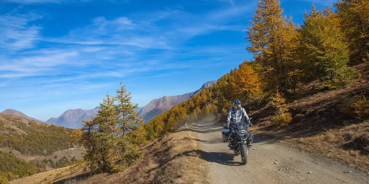 Motociclista su strada sterrata, strada colle delle finestre, con larici dorati e montagne sullo sfondo.