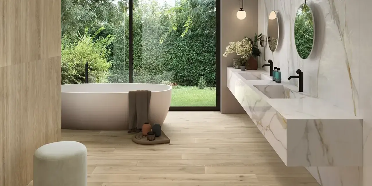 Salle de bain moderne avec baignoire îlot, double vasque en marbre et **pose carrelage imitation parquet** au sol et sur un mur. Vue sur jardin luxuriant.