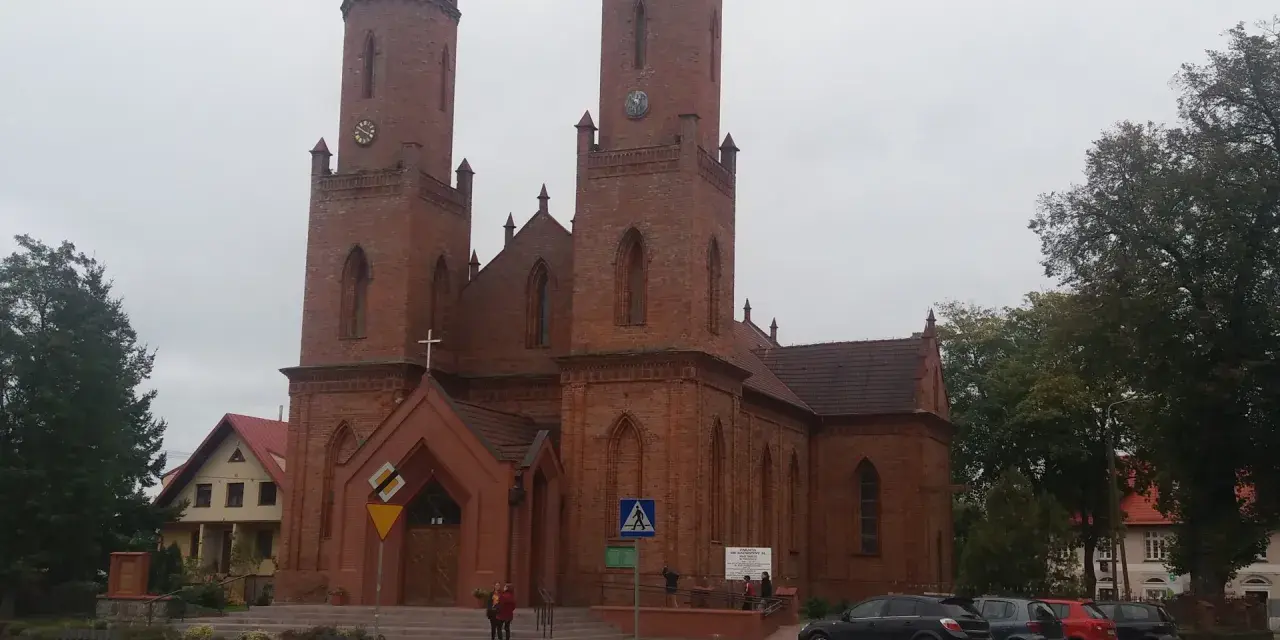 Imponujący kościół w Krokowej z cegły, z dwiema wieżami i zegarami, otoczony zielenią i budynkami.