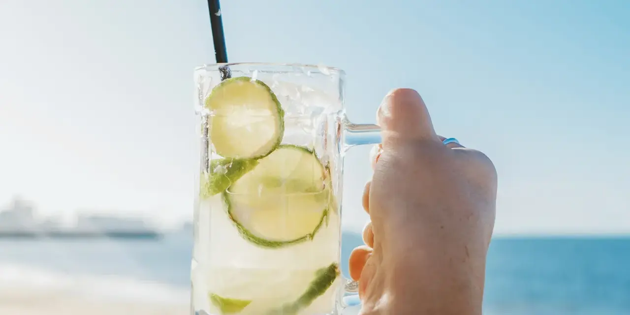 Orzeźwiająca Caipirinha czy Caipiroska z limonką i lodem, trzymana na tle plaży i błękitnego nieba. Idealny napój na wakacje.