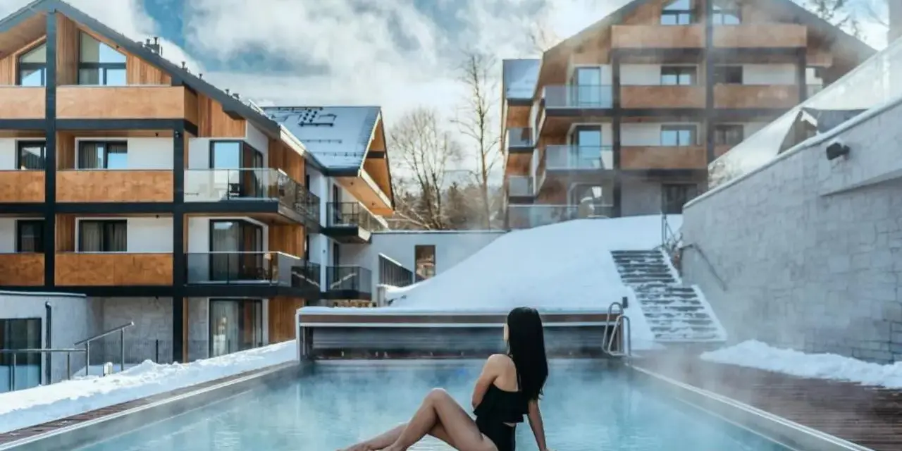 Kobieta w czarnym stroju kąpielowym siedzi przy podgrzewanym basenie. W tle nowoczesny hotel Karpacz w Karpaczu, otoczony śniegiem.