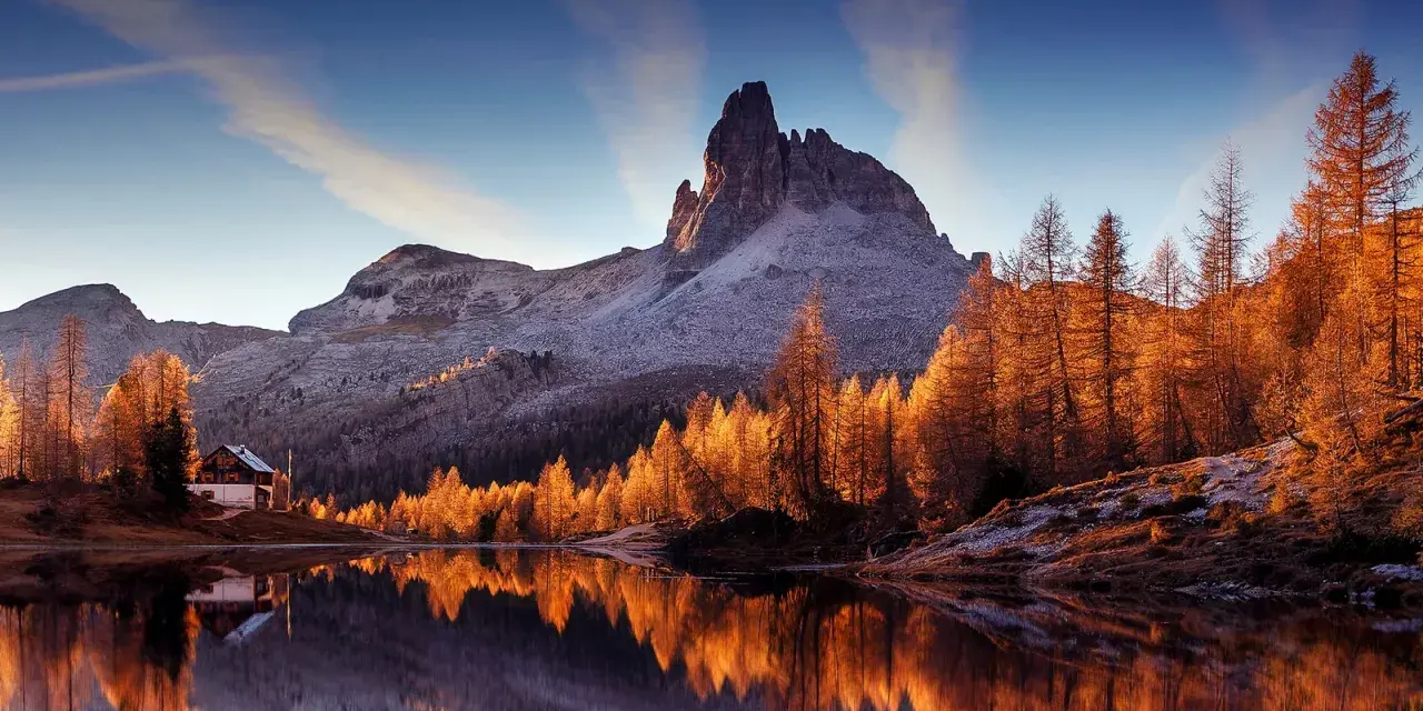 Riflessi dorati sull'acqua di un lago alpino, con un rifugio e montagne innevate. Perfetto per le camminate a Cortina d'Ampezzo.