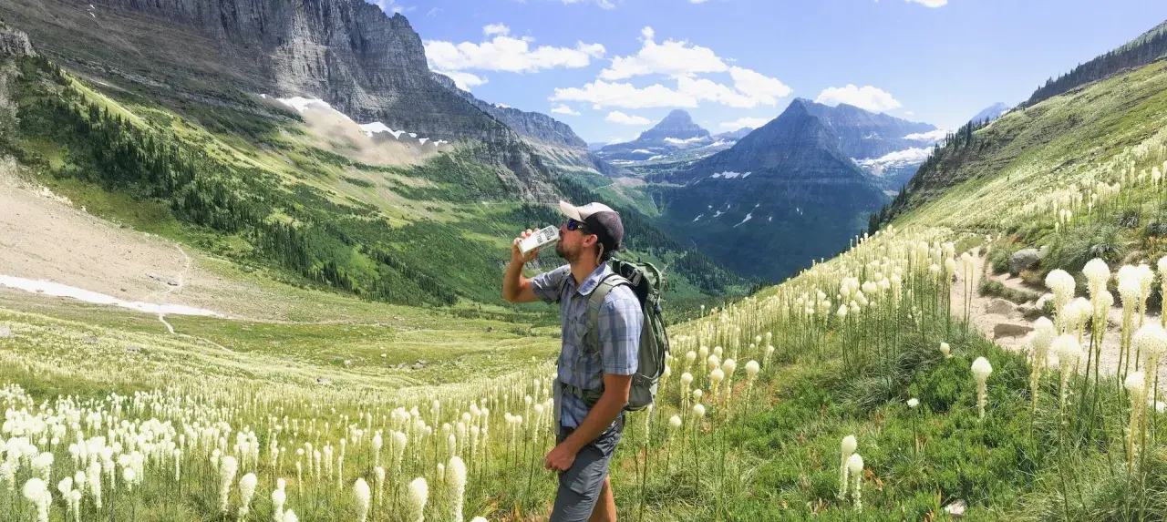 Randonneur buvant de l'eau au milieu d'un champ de fleurs blanches, avec des montagnes majestueuses en arrière-plan. Il espère que cela soulagera sa crampe cuisse.