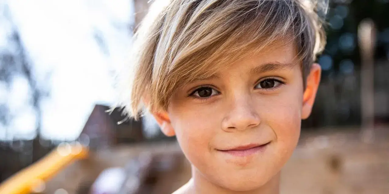 Ein Junge mit blonden Haaren, die nach vorne fallen, lächelt in die Kamera.