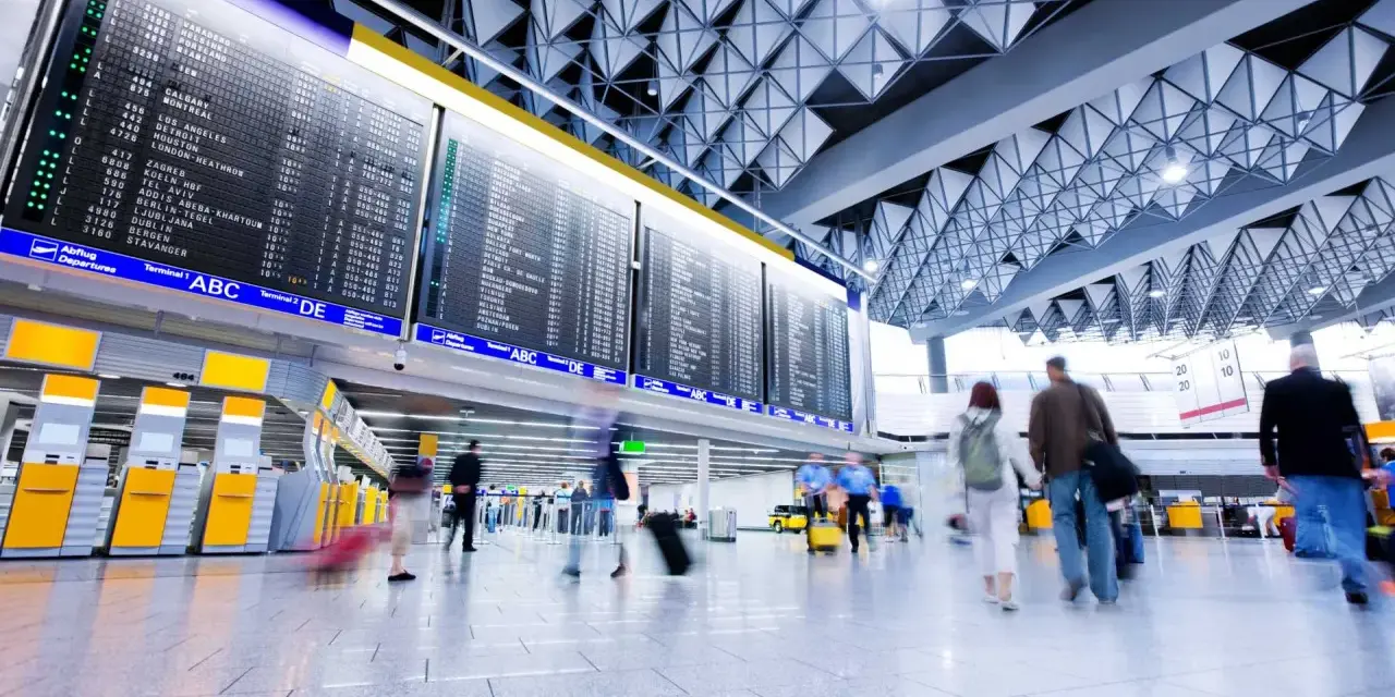 Tablica odlotów na lotnisku, ludzie z bagażami spieszą na boarding.
