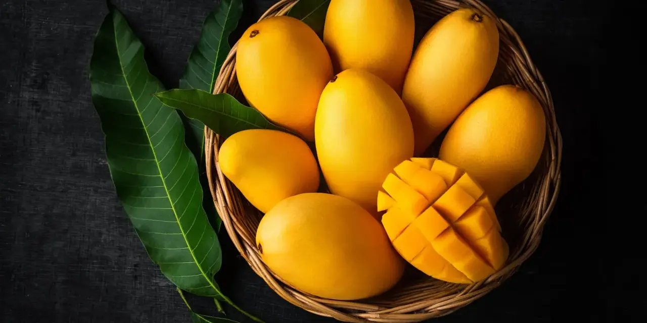 Koszyk pełen soczystych, żółtych mango, jedno przekrojone w kostkę. Obok zielone liście mango.
