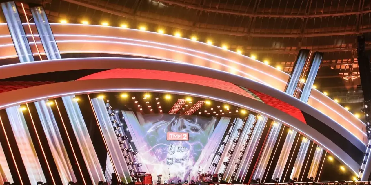 Wielka scena z neonami i ekranami LED, gotowa na Sylwester z Dwójką. Tłum ludzi czeka na rozpoczęcie koncertu.