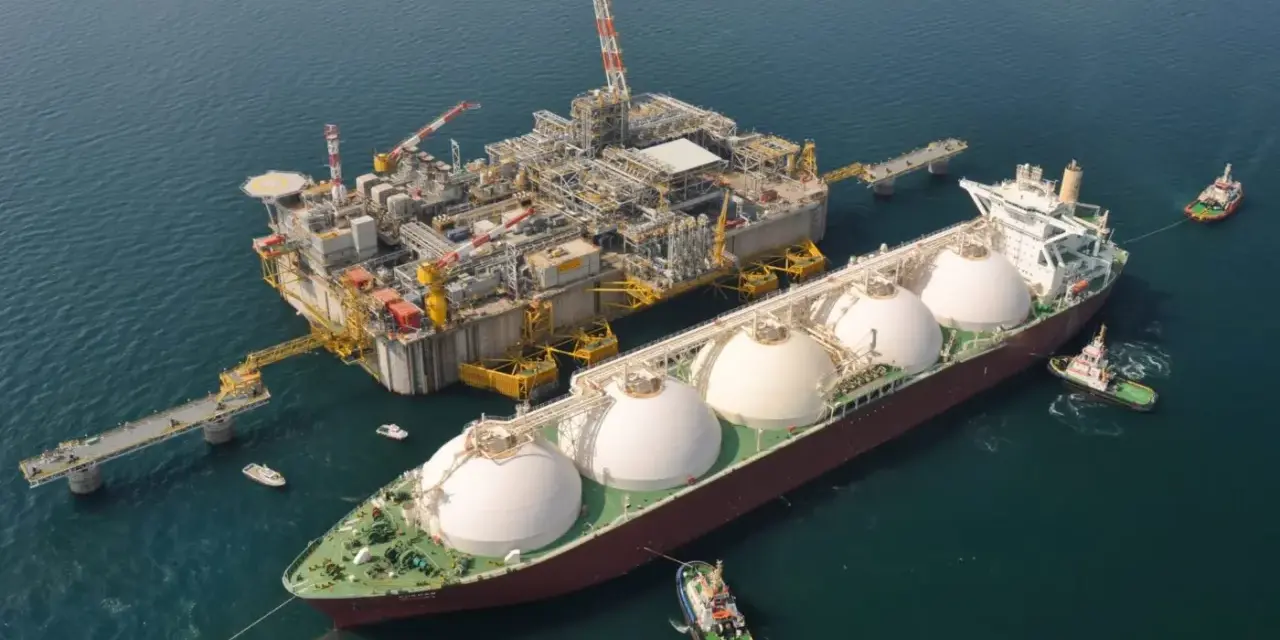 Statek LNG przycumowany do platformy wiertniczej na morzu, holowany przez holowniki.