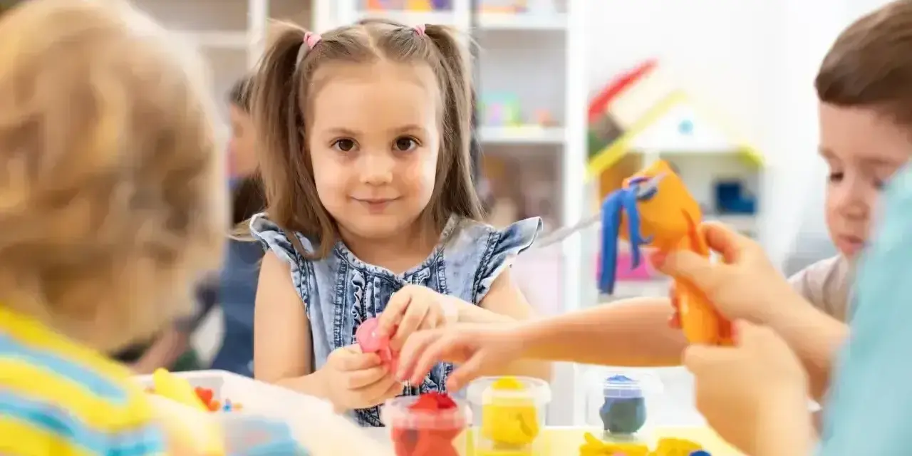 Uśmiechnięta dziewczynka z kitkami bawi się plasteliną, zastanawiając się, w jakim wieku do szkoły pójdzie. Dzieci tworzą kolorowe figurki.