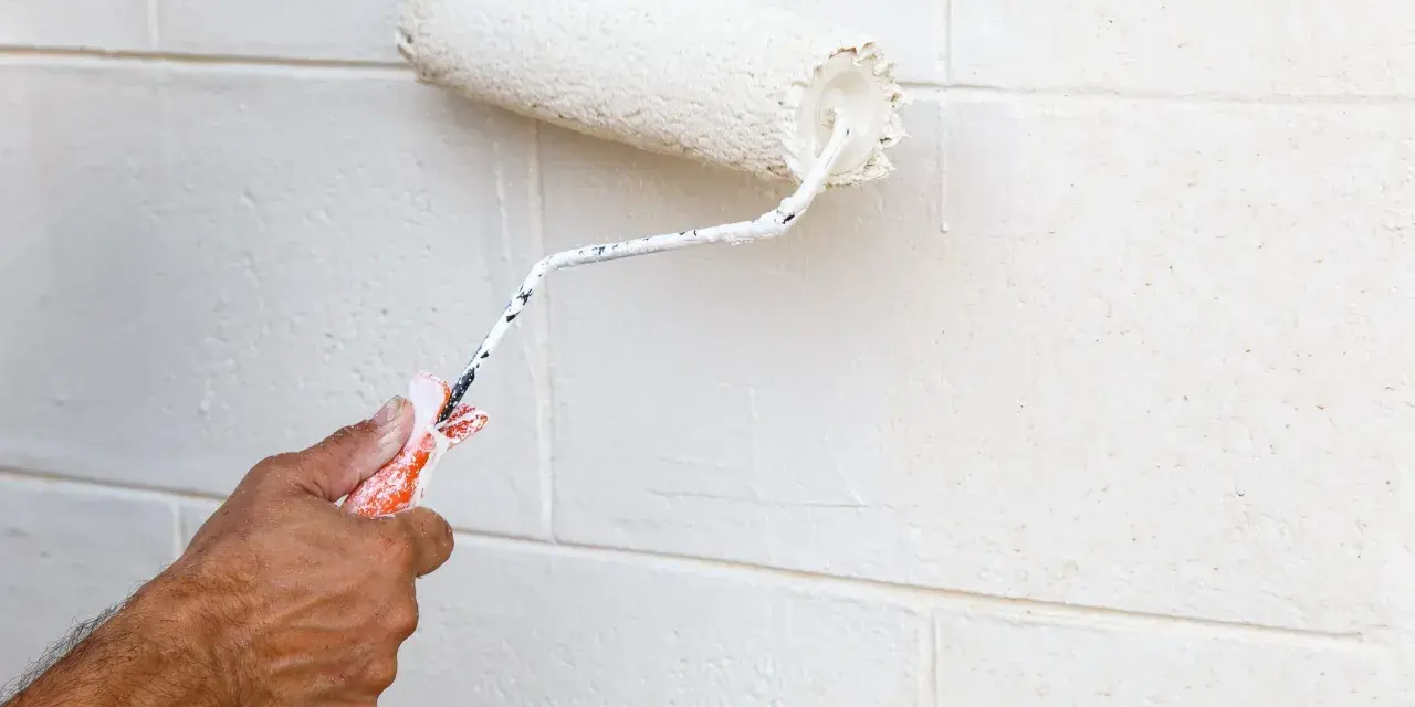 Main tenant, avis peinture carrelage sol : un rouleau blanc applique une nouvelle couche sur un mur en parpaings.