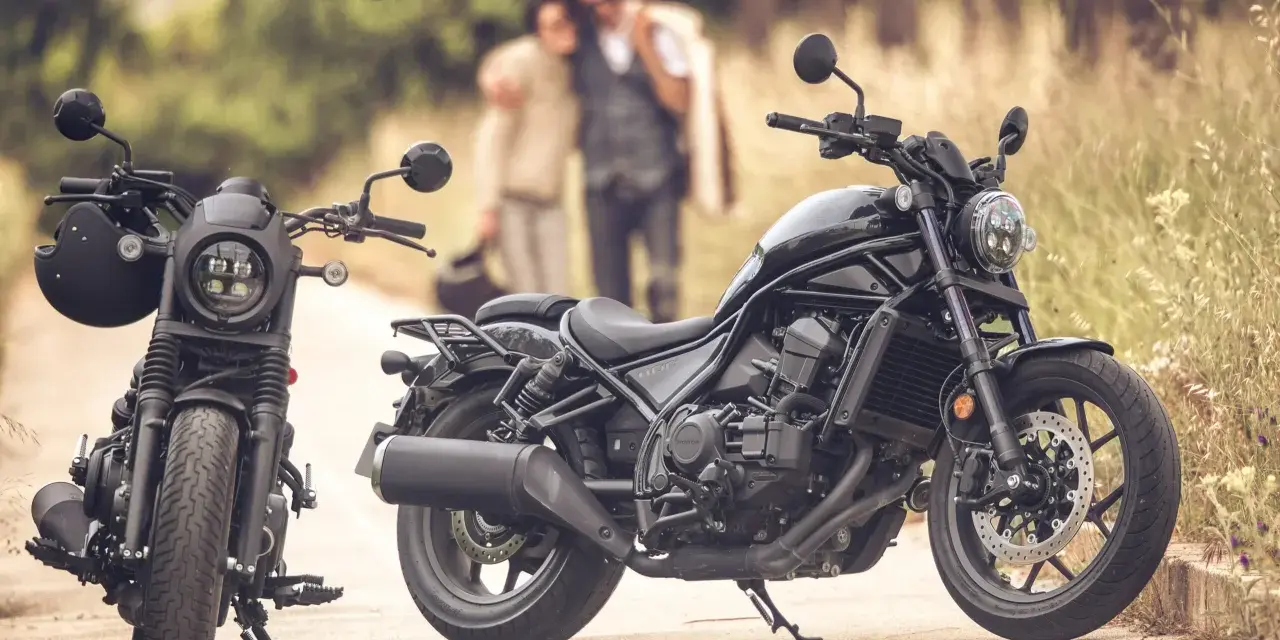 Dos motos negras, una con casco, listas para un viaje. Una pareja abraza en el fondo, disfrutando del paisaje. Una moto sin marchas, perfecta para la aventura.