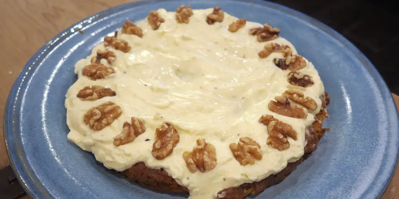 En saftig glutenfri morotskaka med krämig frosting och valnötter på en blå tallrik.