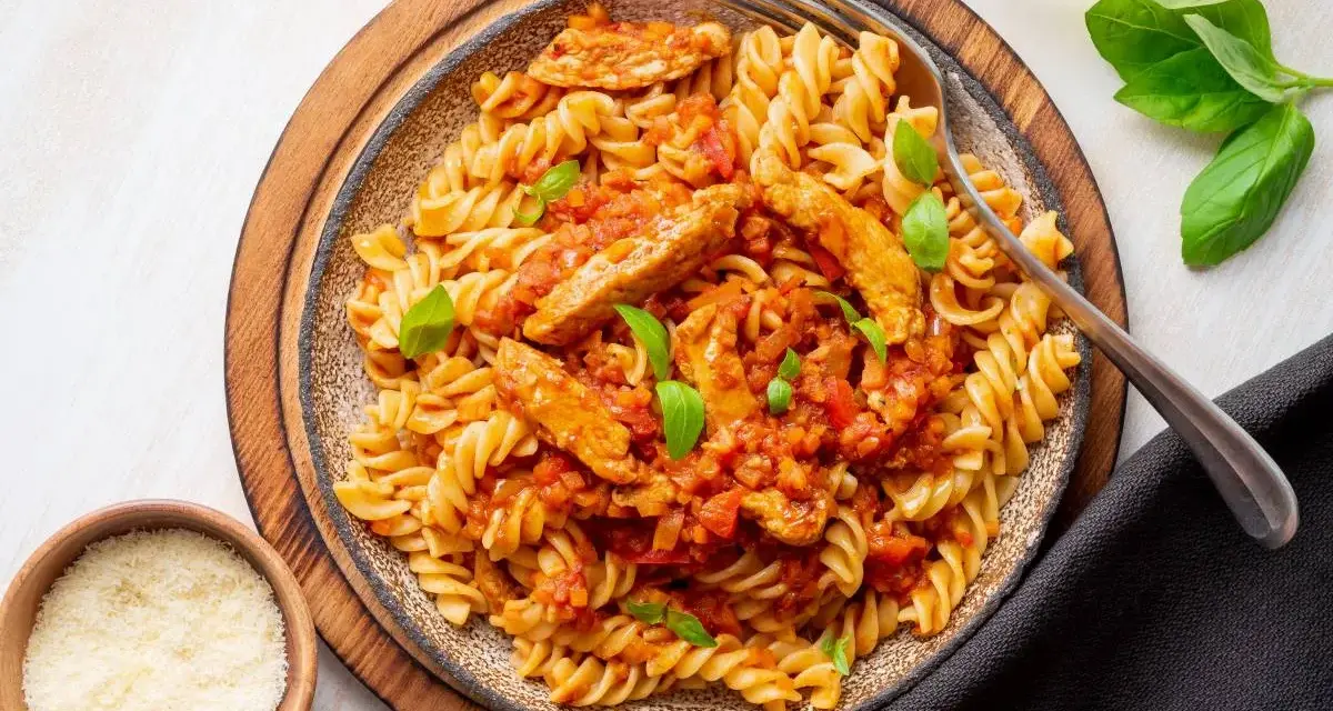 Pyszny makaron z kurczakiem w sosie pomidorowym, udekorowany bazylią. Obok miseczki z parmezanem i ziołami.