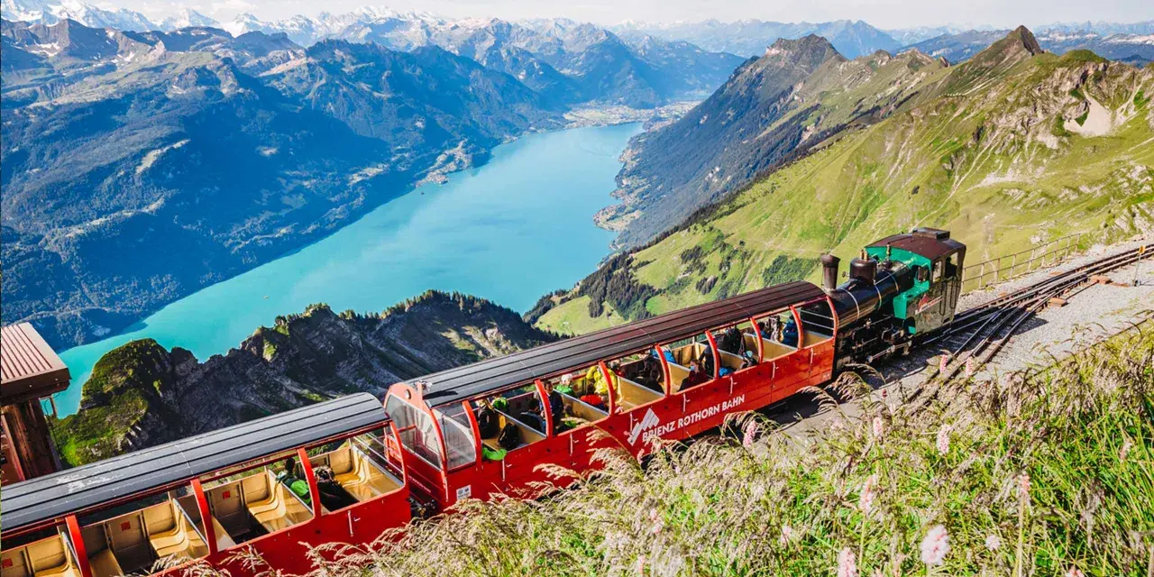 The Brienzer Rothorn train ascends a steep mountain track, offering passengers breathtaking views of a turquoise lake and snow-capped peaks.