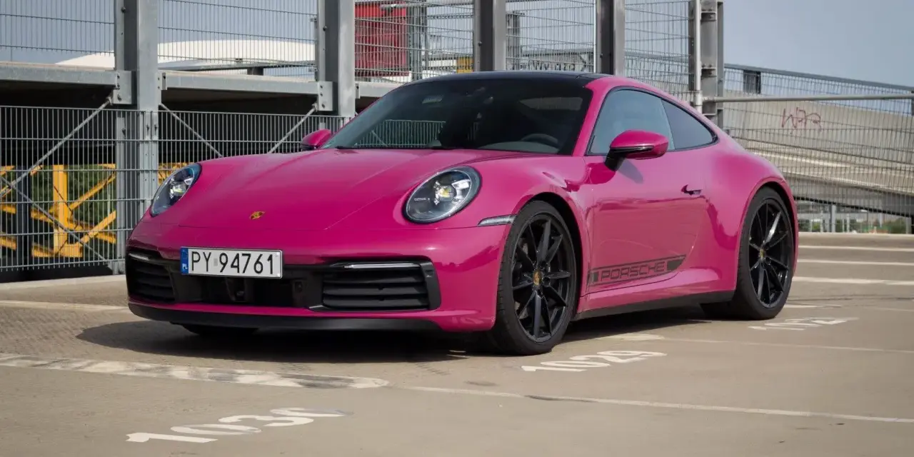 Różowy Porsche 911 Carrera 4S z czarnymi felgami i naklejką PORSCHE na progu, zaparkowany na zewnątrz.