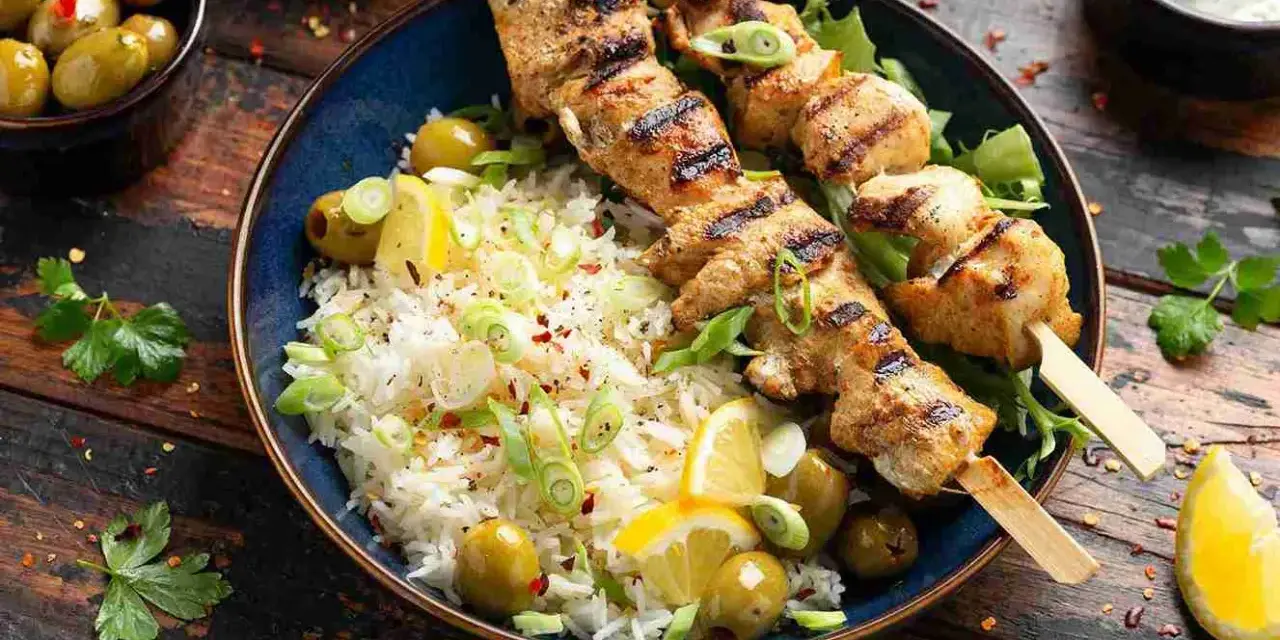 Deliciosos pinchitos de pollo a la parrilla servidos con arroz blanco, aceitunas verdes, rodajas de limón y cebolleta picada.
