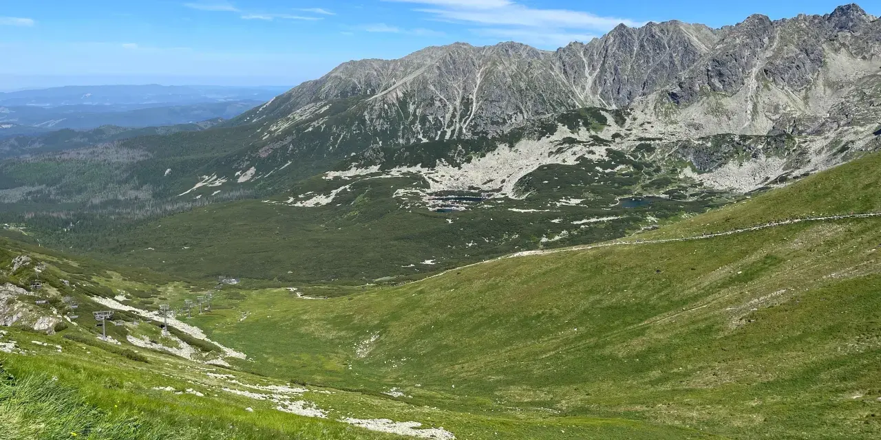 Widok na Tatry z zielonymi zboczami i skalistymi szczytami. W oddali widać budynki. Zastanawiasz się, ile km jest na Kasprowy Wierch?