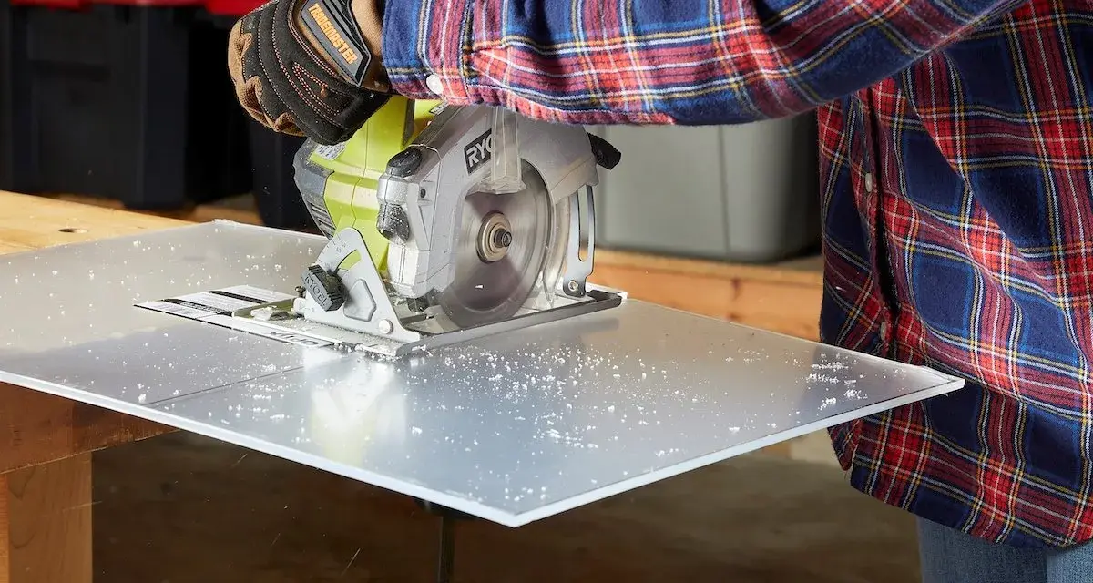 A person in a plaid shirt and gloves uses a circular saw to cut a sheet of plexiglass, creating small plastic shavings. This demonstrates that plexiglass is easy to cut with the right tools.