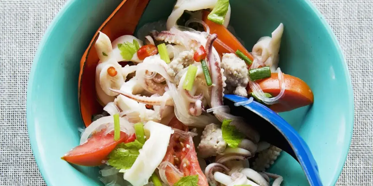 Ensalada tailandesa con fideos de cristal, calamares, tomate y cebolla, servida en un cuenco azul. El plato de sepia en salsa se ve fresco y apetitoso.