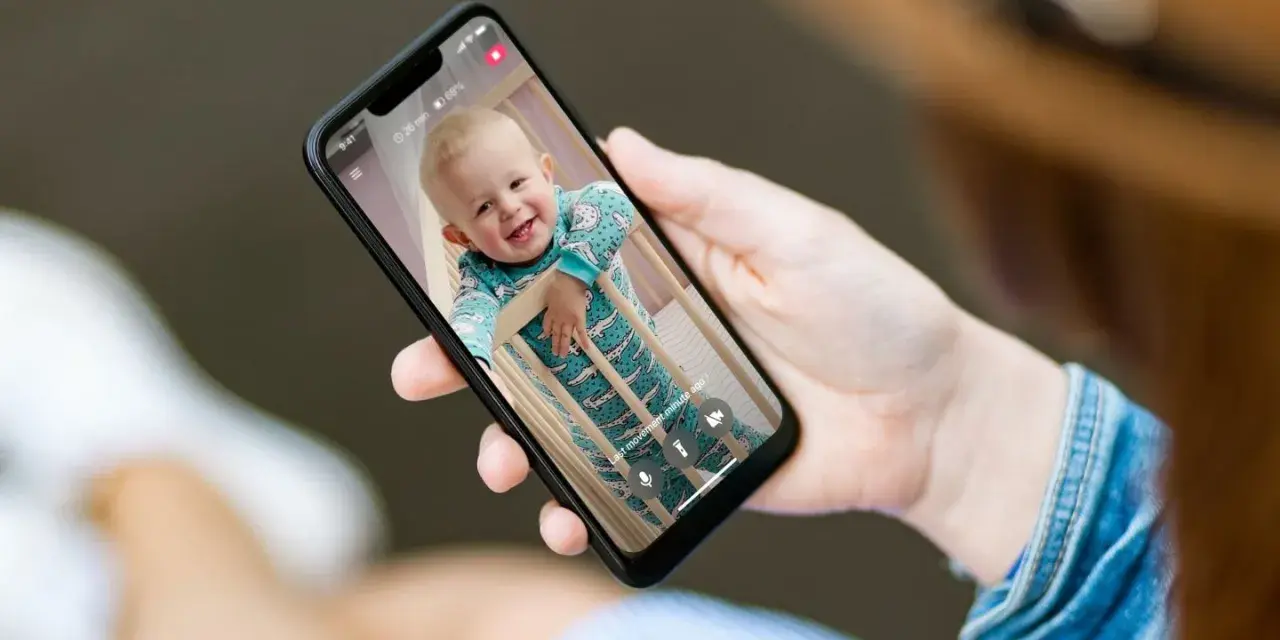 Ein lächelndes Baby im Strampler schaut aus dem Gitterbett. Eine Hand hält das Handy als Babyphone, um das Kind im Blick zu behalten.
