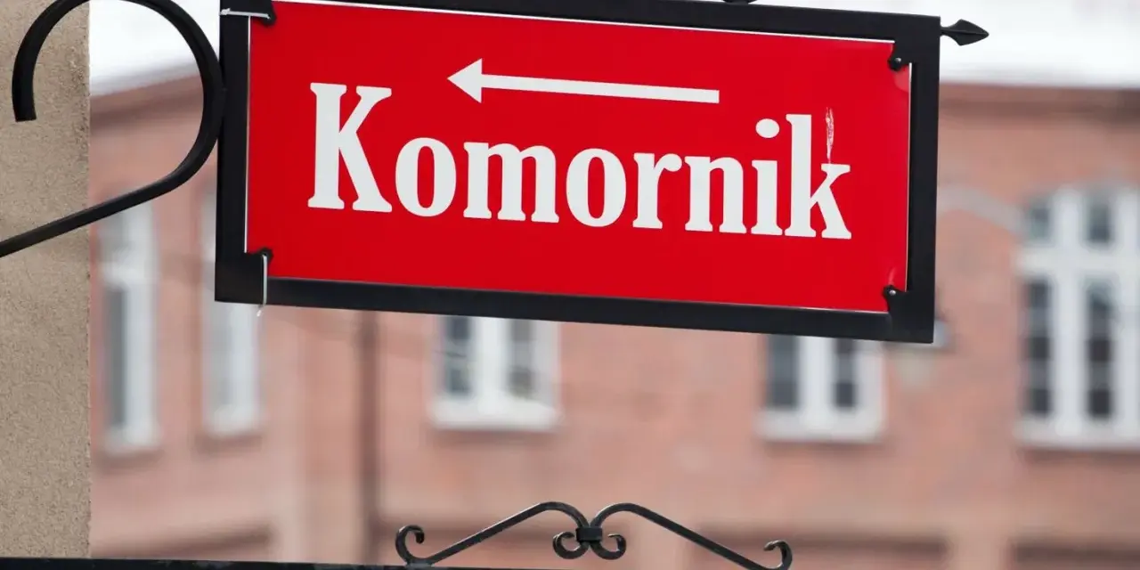 Czerwona tablica z napisem "Komornik" i strzałką w lewo. W tle budynek, może to być miejsce, gdzie zasiłek chorobowy jest zagrożony przez komornika.