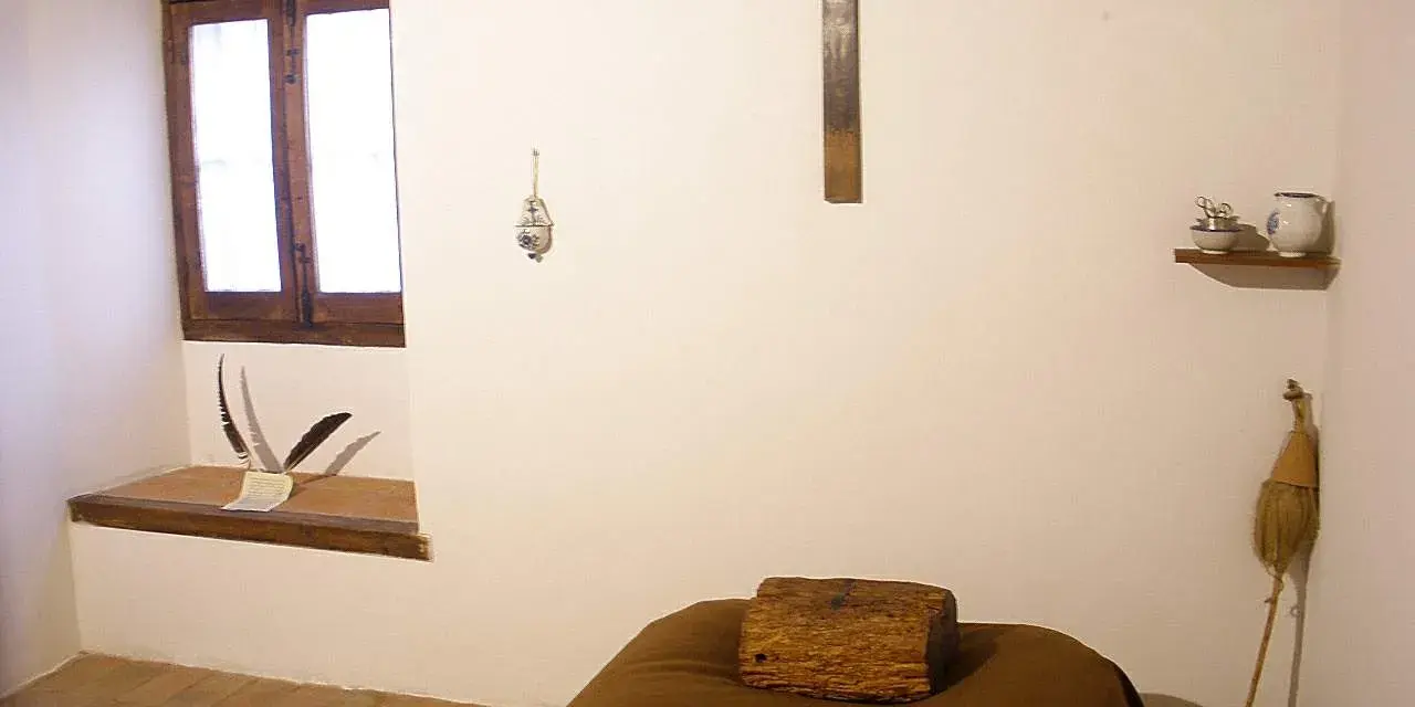 A monk's simple cell, showing where monks sleep: a narrow bed with a wooden block for a pillow, a crucifix, and a window.