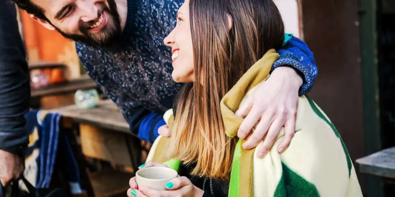 Pareja sonriente compartiendo una manta y una taza de té. Él le abraza por detrás. Ella le dice "muchas gracias en francés".