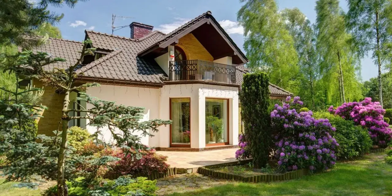 Dom z balkonem i tarasem, otoczony bujną zielenią i kwitnącymi rododendronami. Występ ściany podkreśla nowoczesny charakter budynku.