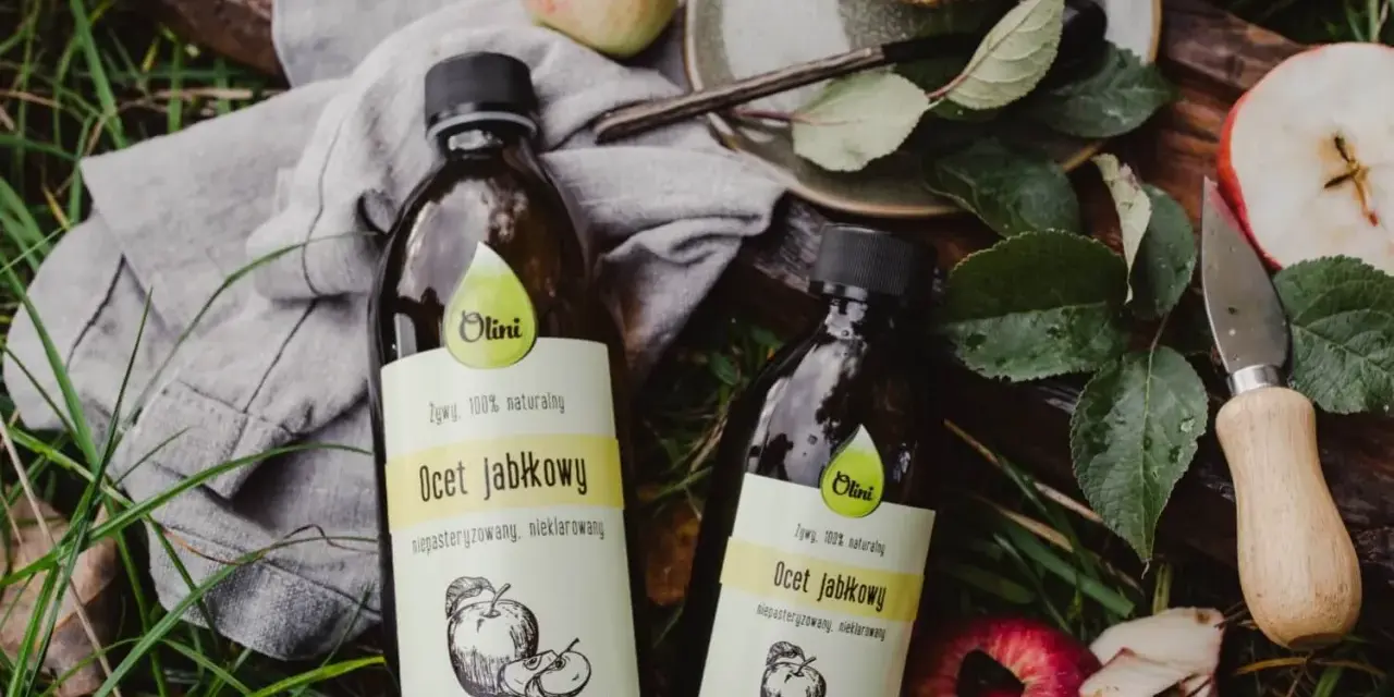 Dwie butelki naturalnego octu jabłkowego Olini, idealnego dla dzieci, leżą wśród jabłek i liści.