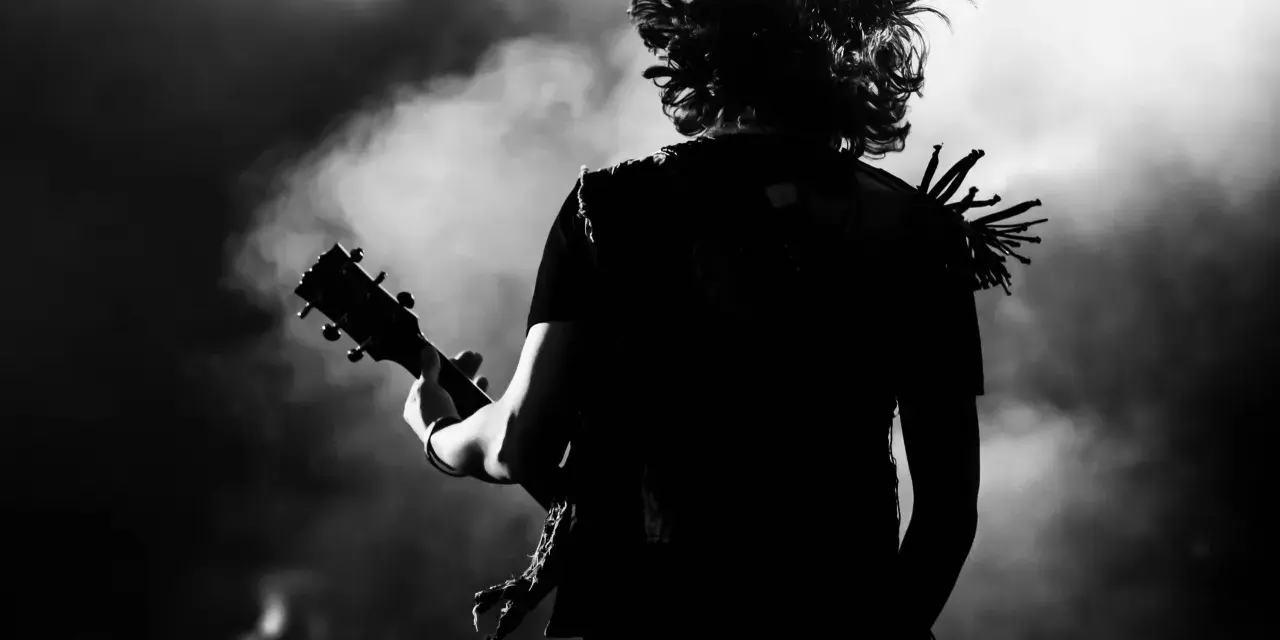 Muzyk z rozwianymi włosami gra na gitarze, w dymie i świetle sceny. Energia gatunku rockowego w czerni i bieli.