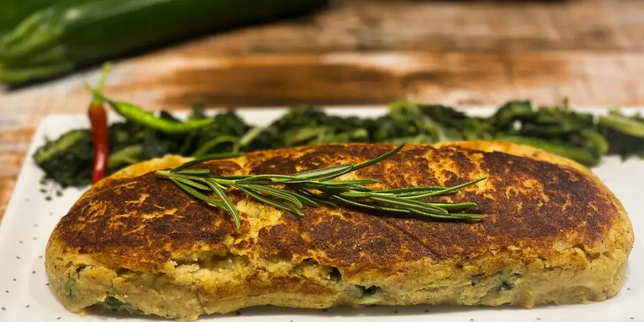 Polpettone di verdure dorato, guarnito con rosmarino, servito con contorno di verdure verdi e peperoncino.