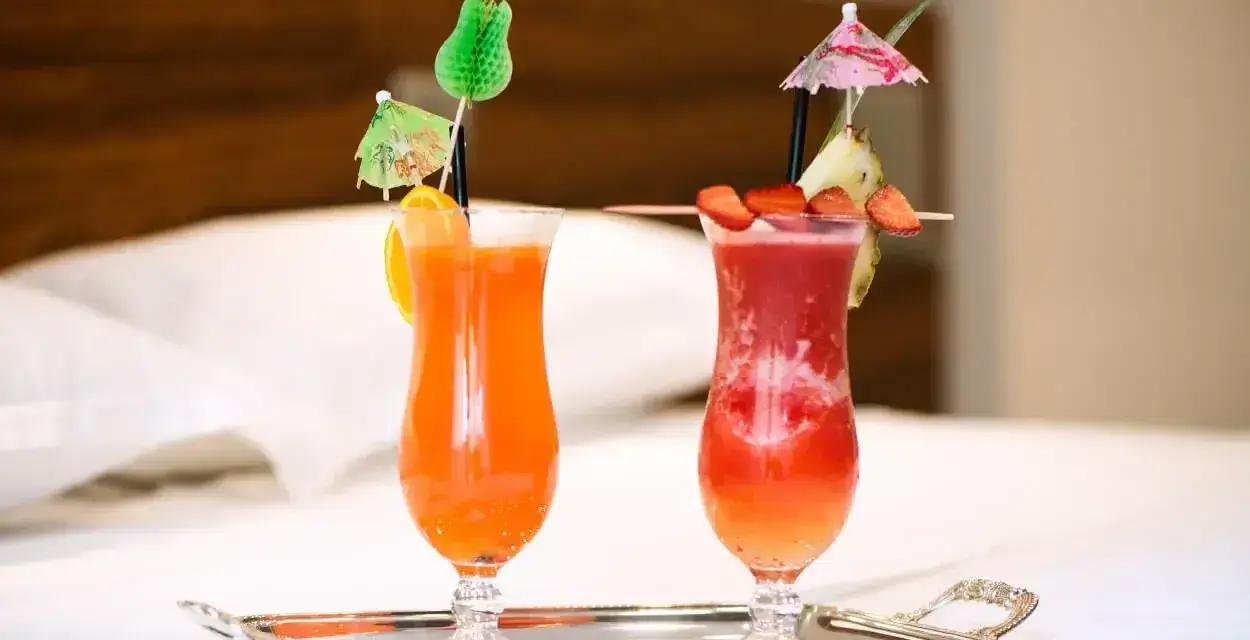 Dwa orzeźwiające drinki: jeden pomarańczowy z pomarańczą, drugi czerwony z truskawkami i ananasem, oba z parasolkami. Idealne na wakacje, może z dodatkiem triple sec.