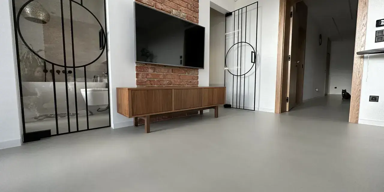 Nowoczesne wnętrze z betonową podłogą, co to jest mikrocement, która nadaje mu surowego charakteru. W tle widać łazienkę i salon.