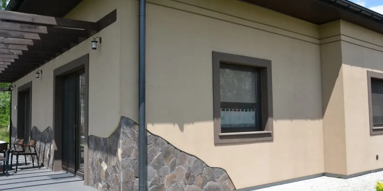Elewacja domu w kolorze kawa z mlekiem, ozdobiona kamiennym cokołem. Widoczne okna i drzwi tarasowe.