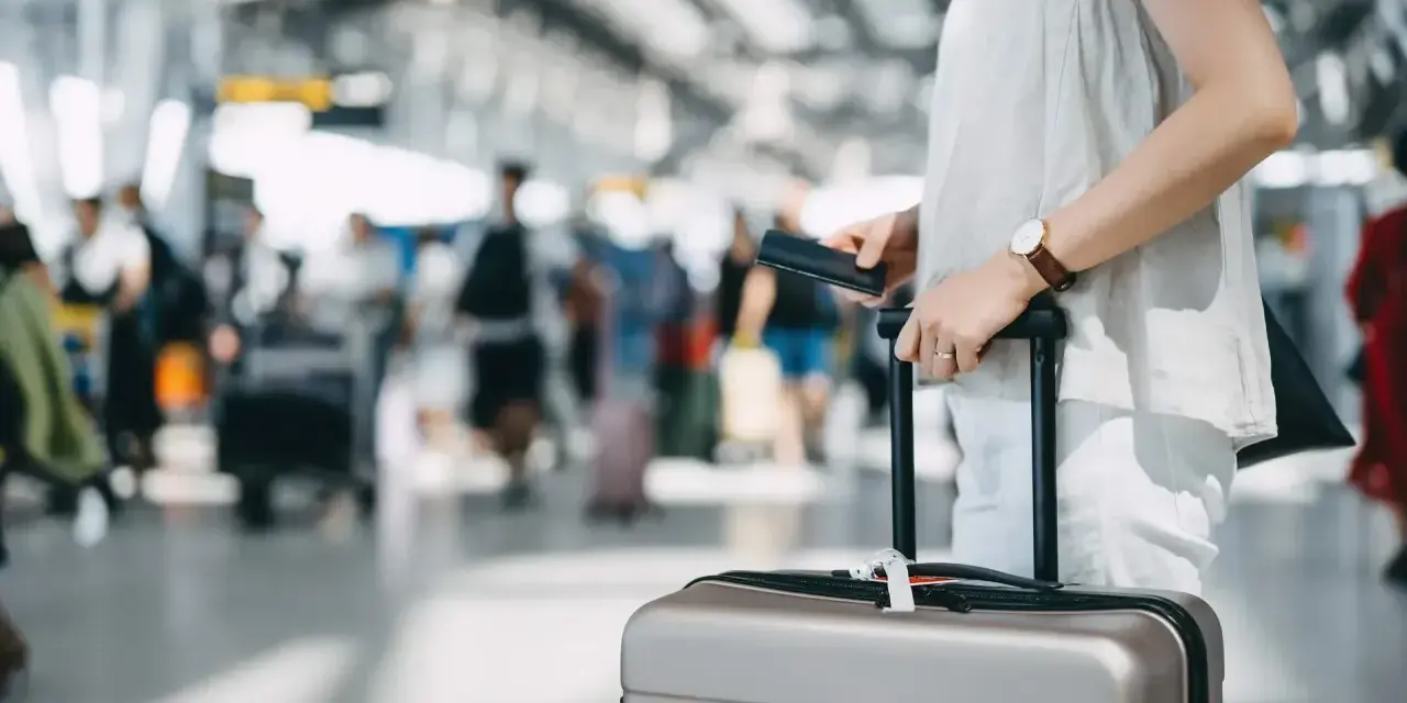 A traveler holds a passport and suitcase, mindful of carry-on size limits in a busy airport.