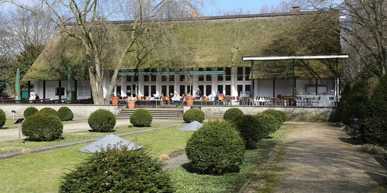 Das Teehaus im Englischen Garten Berlin mit seinem Reetdach und der Terrasse, auf der Gäste sitzen, ist von gepflegten Rasenflächen und Bäumen umgeben.