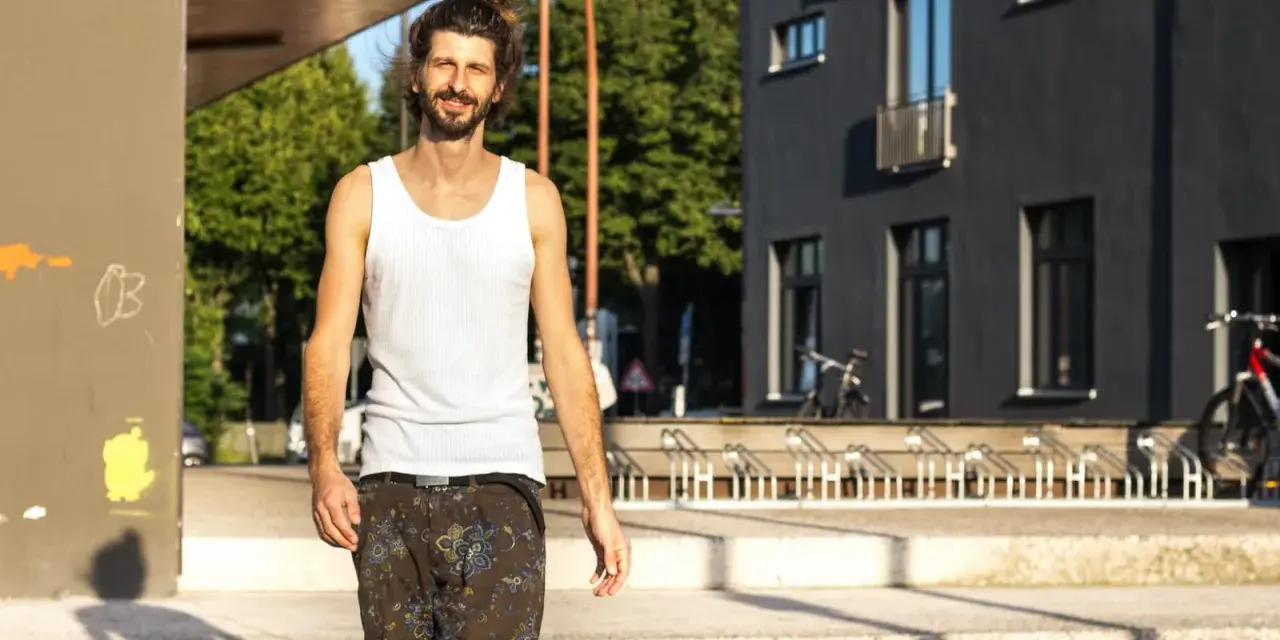 Uśmiechnięty mężczyzna w białej podkoszulce i wzorzystych spodenkach, z włosami związanymi w kok, idzie ulicą. Wygląda jakby właśnie skończył trening, a jego styl przypomina "żonobijka".