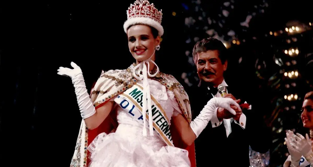 Uśmiechnięta Miss International w koronie i białej sukni, z szarfą "Miss Polonia". Obok niej mężczyzna w smokingu.